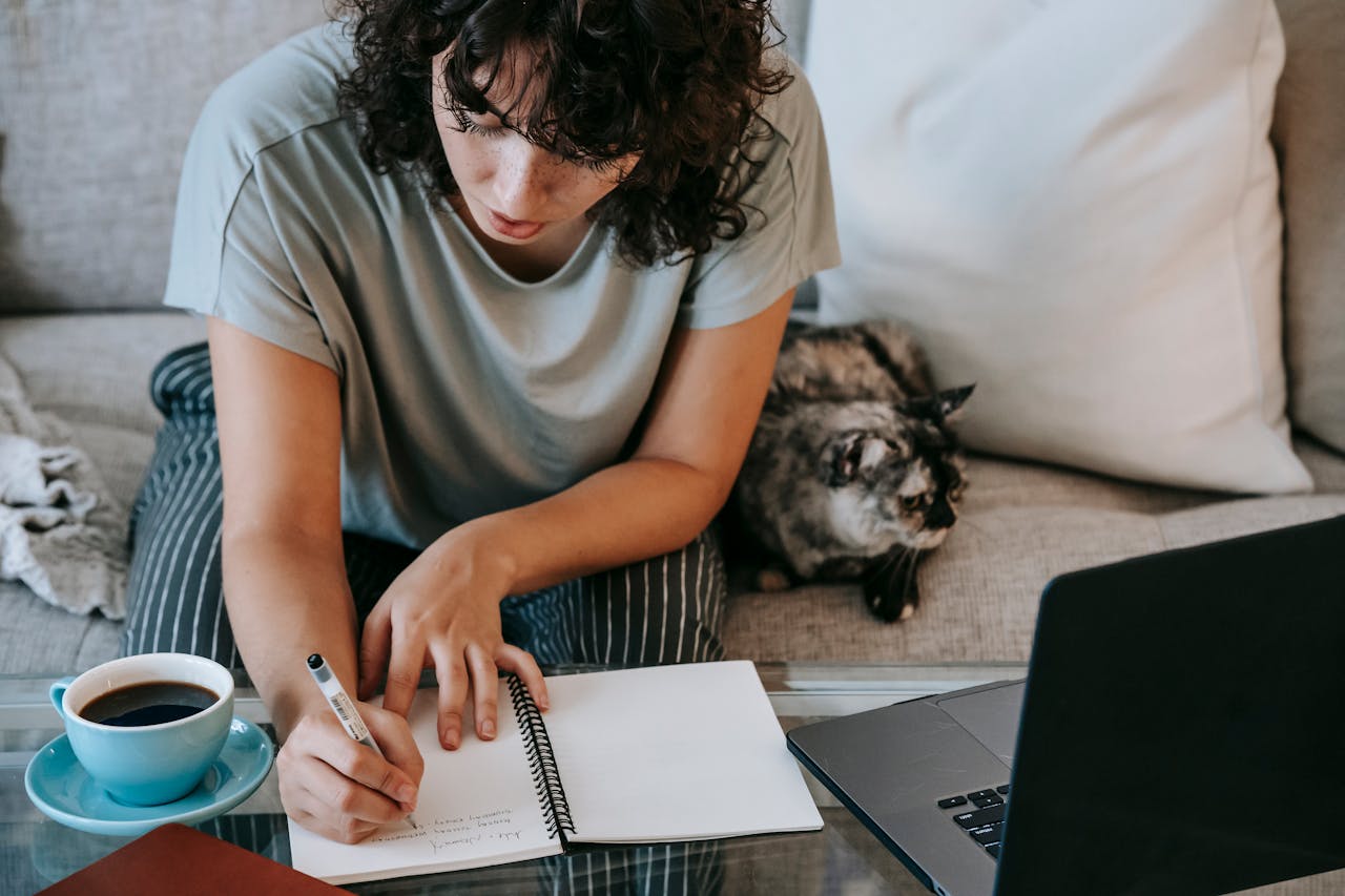 Young woman studying at home with a laptop, taking notes on a notebook, accompanied by a cat on the sofa.